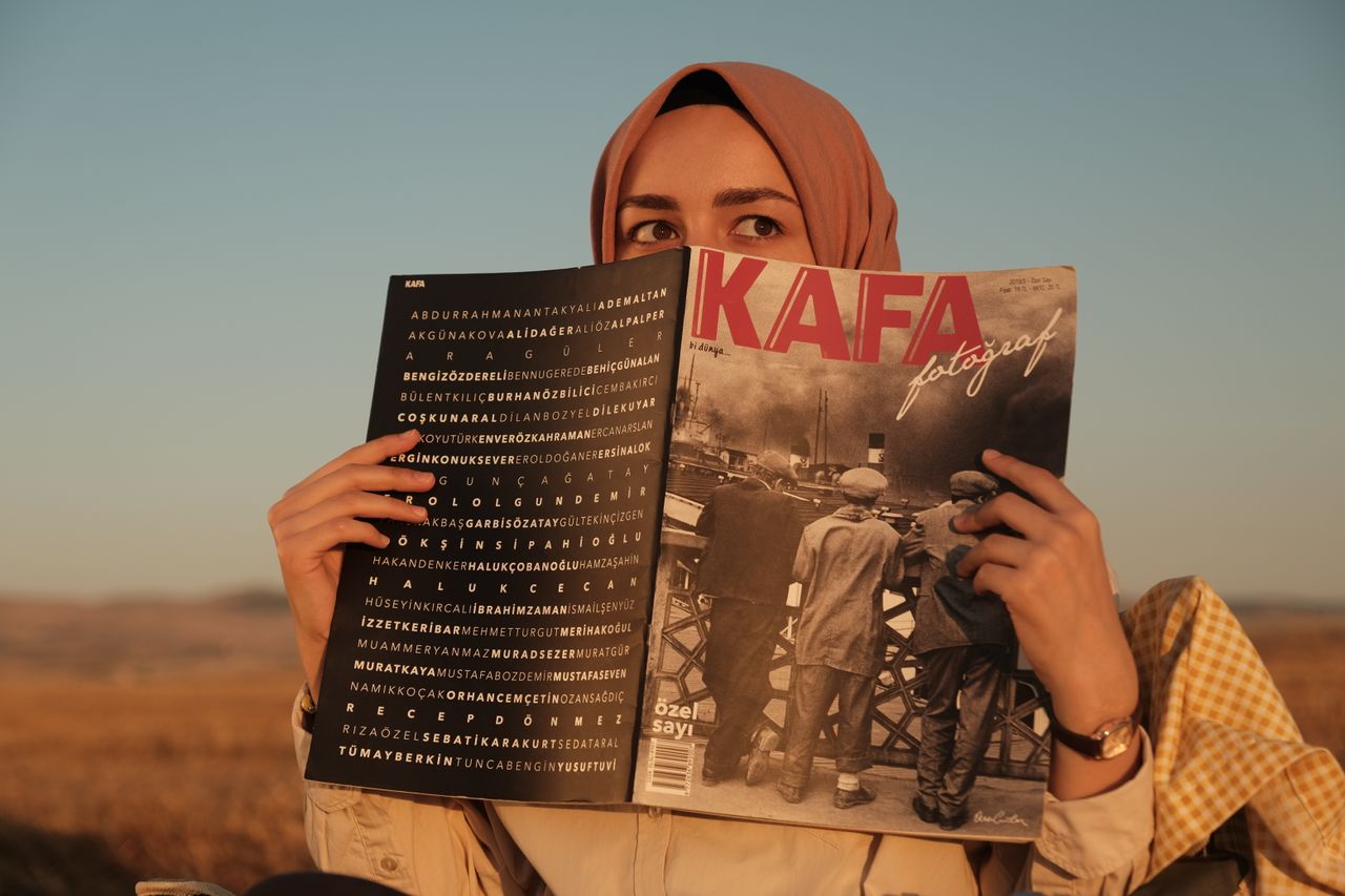 A woman in a hijab holds a magazine while enjoying the sunset outdoors.