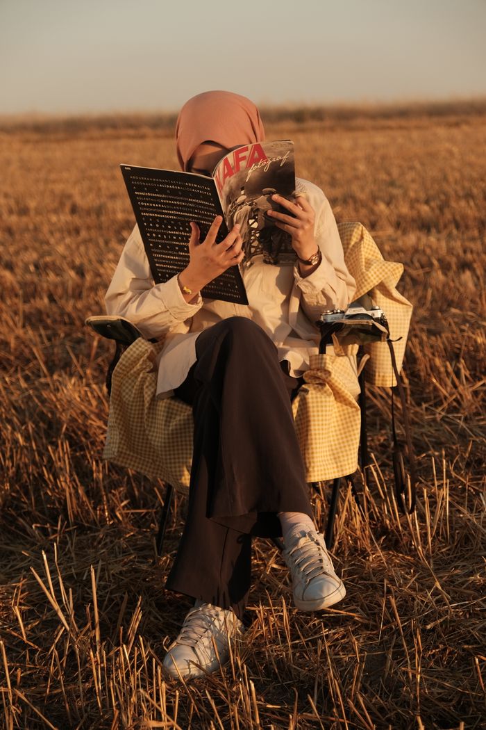 services-02 A woman in a hijab sits in a field reading a magazine, enjoying the peaceful sunset.